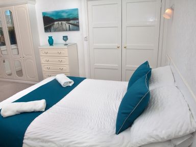 A bedroom with a double bed with teal pillows and runner two rolled white towels dresser with two blue vases wardrobe with mirrored doors painting of a pier double-door closet Weymouth Bay Apartment B Greenhill