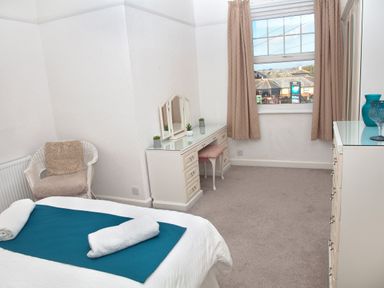 A bedroom with a bed, a white wicker chair, a vanity desk with a stool, a chest of drawers, and a window with beige curtains at Weymouth Bay Apartment B in Greenhill