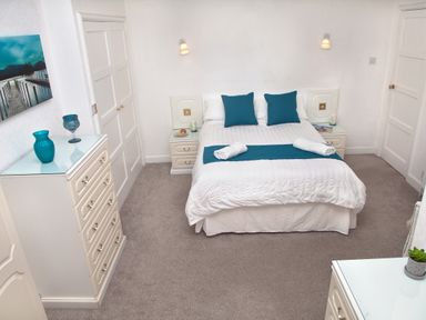 A bedroom with a double bed with blue pillows and white bedding with dressers and a painting on the wall at Weymouth Bay Apartment B in Greenhill