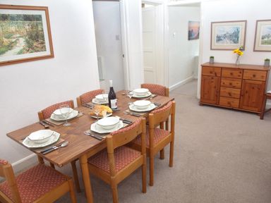 A dining room with a wooden table set for six with plates glasses and a bottle of wine and a wooden sideboard with framed pictures in Weymouth Bay Apartment B Greenhill