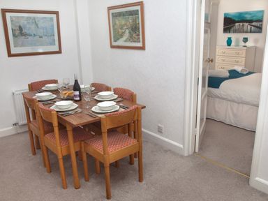 A dining area with a wooden table set for six and a glimpse of a bedroom with a bed and dresser at Weymouth Bay Apartment B in Greenhill