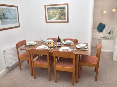 A dining table with six chairs set with plates glasses cutlery and a bottle of wine in a white room at Weymouth Bay Apartment B in Greenhill