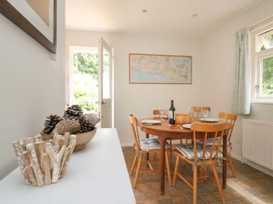 A dining room with a round wooden table set with plates and glasses and wooden chairs with cushions at Woodlands Cottage in Ringstead