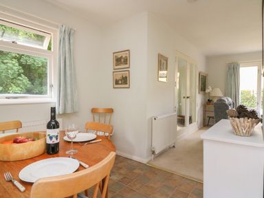 A dining area with a wooden table set with plates utensils a bottle of wine and a bowl of fruit next to a window with curtains that opens onto a living room with seating and decorated with pine cones at Woodlands Cottage Ringstead