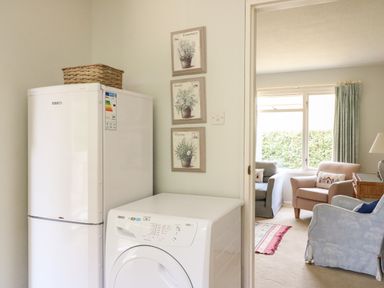 A kitchen area with a white fridge freezer and washing machine next to an entrance leading to a sitting room with chairs and a window at Woodlands Cottage in Ringstead