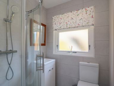 A bathroom with a shower enclosure, a toilet, a window with a floral blind, a small white cabinet, and a wooden framed mirror at Woodlands Cottage in Ringstead