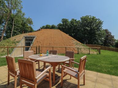 An outdoor patio with a wooden table and six chairs near a mound covered with soil and grass at Woodlands Cottage in Ringstead