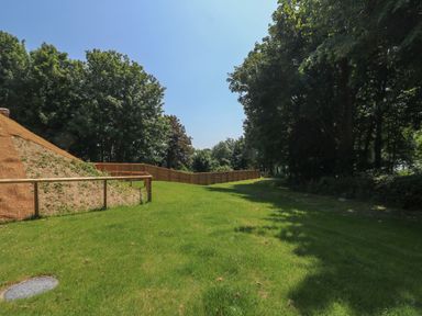 A grassy open area with trees, a wooden fence, and a dirt slope at Woodlands Cottage in Ringstead