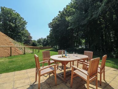 An outdoor patio with a round wooden table and six wooden chairs on tiled flooring surrounded by grass and trees at Woodlands Cottage in Ringstead