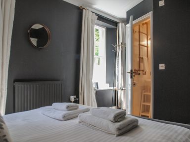 A bedroom with a bed folded towels a open door leading to a bathroom a window with curtains and a round mirror on the wall at 16 Courtenay Street Salcombe