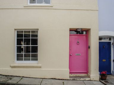 A cream colored building with a pink door and a white framed window beside it with a blue door and potted plant nearby at 16 Courtenay Street in Salcombe