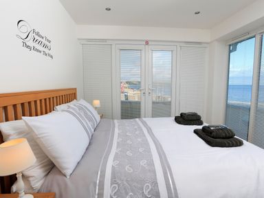 A bedroom with a double bed with gray and white bedding folded towels on the bed two bedside lamps and windows with blinds overlooking the sea at 1 At The Beach in Torcross