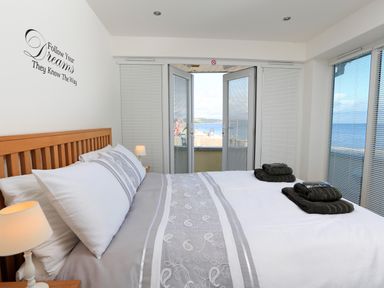A bedroom with a double bed with gray and white bedding and folded towels on it and a view of the sea through glass doors at 1 At The Beach in Torcross