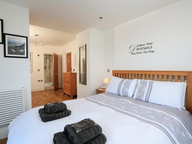 A bedroom with a bed, wooden furniture, towels on the bed, wall art, and a view into an attached bathroom at 1 At The Beach in Torcross