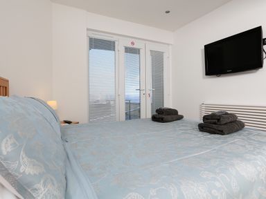 A bedroom with a bed covered with blue bedding and folded towels a wall-mounted tv and glass doors with blinds at 1 At The Beach in Torcross