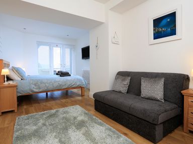 A bedroom with a bed bedside tables a gray couch two patterned cushions a rug and a wall-mounted television at 1 At The Beach in Torcross