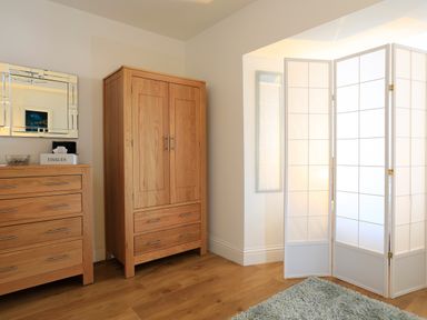 A room with wooden chest of drawers and wardrobe next to a white folding room divider and a green rug on a wooden floor at 1 At The Beach in Torcross