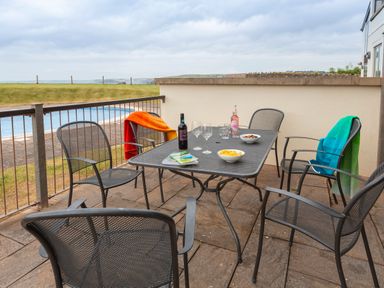 An outdoor patio with a metal table and six chairs with drinks bowls and blankets near a body of water at 2 Ocean's Edge in Thurlestone