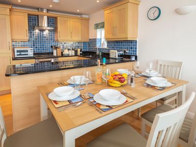 A kitchen with wooden cabinets blue tiled backsplash and a dining table set for six with white dishes and fruit bowl at 2 Oceans Edge Thurlestone