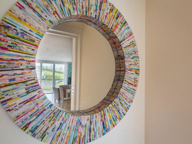 A round colorful framed mirror reflecting a room with chairs and large windows at 2 Oceans Edge in Thurlestone