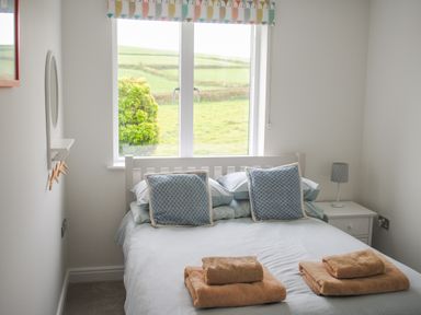 A bedroom with a bed with blue pillows and folded towels near a window with a patterned valance at 2 Oceans Edge in Thurlestone