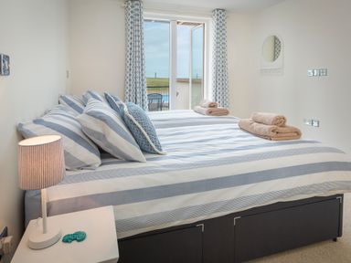 A bedroom with a bed with striped bedding and towels on top a bedside table with a lamp and a door leading outside at 2 Oceans Edge in Thurlestone