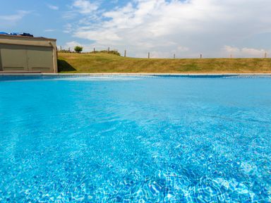 An outdoor swimming pool with clear blue water next to a grassy area and partly cloudy sky at 2 Ocean's Edge in Thurlestone