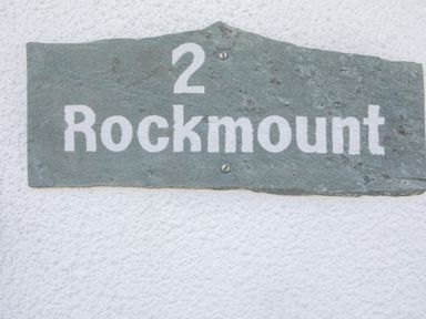 A rectangular slate house sign with the text 2 Rockmount mounted on a white textured wall at 2 Rockmount Salcombe