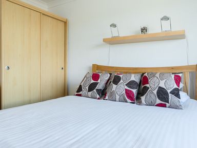 A bedroom with a wooden bed frame white bedding three leaf patterned cushions a wooden shelf with two lamps and a glass vase and a wooden sliding door wardrobe at 2 Rockmount in Salcombe