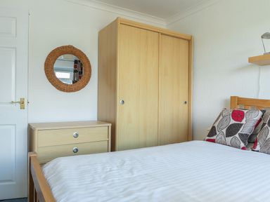 A bedroom with a bed with patterned cushions a wooden wardrobe a chest of drawers and a round wicker mirror on the wall at 2 Rockmount in Salcombe