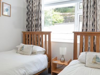 A bedroom with two single wooden beds separated by a wooden nightstand with a lamp and a large window behind at 2 Rockmount in Salcombe