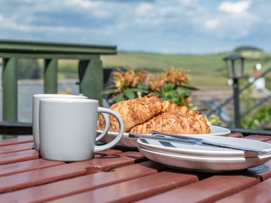 Two white mugs and plates with croissants and cutlery on a wooden table outdoors at 2 Rockmount in Salcombe