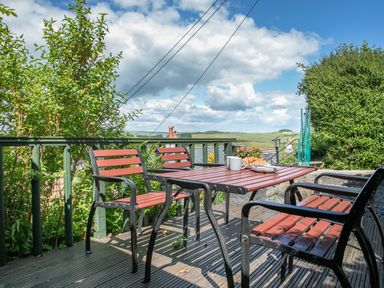 An outdoor wooden deck with a table and chairs surrounded by green plants and a view of the countryside at 2 Rockmount in Salcombe