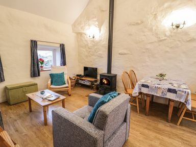 A living room with a gray armchair, wooden coffee table, small dining table with four chairs, wood stove, and a window with curtains at Y Bwthyn in Elidir Fach Mountain near Llanberis