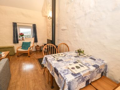 A dining table with placemats and chairs next to a wood stove and a chair by a small window at Y Bwthyn Elidir Fach Mountain near Llanberis