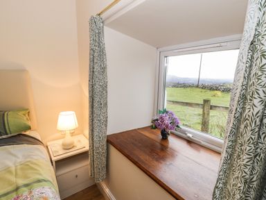 A bedroom corner with a bed next to a nightstand with a lamp and a windowsill with flowers at Y Bwthyn Elidir Fach Mountain near Llanberis
