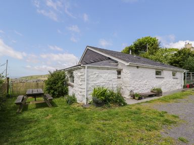 A white stone cottage with a dark roof and a wooden picnic table outside on the grass at Y Bwthyn in Elidir Fach Mountain near Llanberis