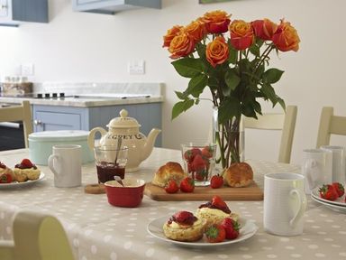 A kitchen table with a teapot jam scones strawberries mugs and a vase of roses at 3 Eddystone Row in Salcombe