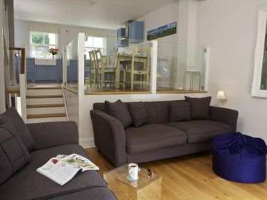 A living room with two brown sofas a magazine on one and a coffee cup on a glass table with a blue bean bag and a dining area with chairs and blue cabinets at 3 Eddystone Row in Salcombe