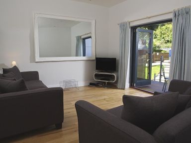 A living room with two dark sofas a large wall mirror a small TV on a stand and an open door leading to a garden at 3 Eddystone Row in Salcombe