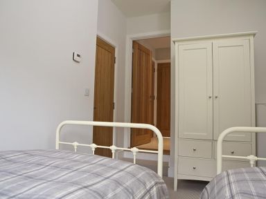 A bedroom with two beds in grey plaid bedding and a white wardrobe near wooden doors at 3 Eddystone Row in Salcombe