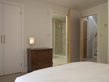 A bedroom with a wooden chest of drawers next to a double wardrobe and two open doors showing a bathroom and a staircase at 3 Eddystone Row in Salcombe