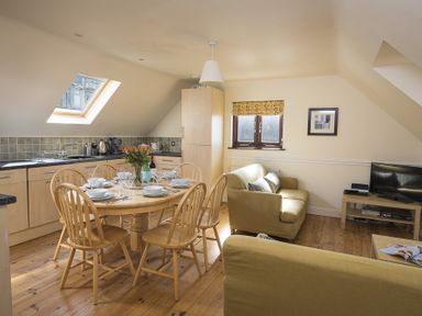A kitchen dining area with a round wooden table set for six people and two sofas by a window at 4 Armada House in Hope Cove