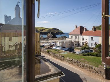 View from an open window showing a street with parked cars houses a stone wall and boats near the sea at 4 Armada House in Hope Cove