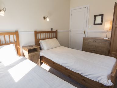 A bedroom with two single beds a wooden nightstand a chest of drawers a lamp a mirror and a closed door at 4 Armada House in Hope Cove