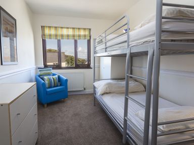 A bedroom with a bunk bed, a blue armchair, a chest of drawers, and a window with striped blinds at 4 Armada House in Hope Cove