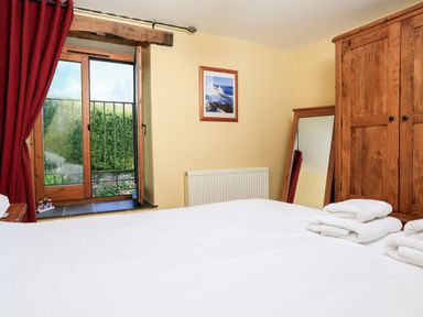A bedroom with a large bed folded towels a wooden wardrobe a standing mirror and an open window with red curtains at Cowslip Cottage in Frogmore