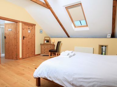 A bedroom with a wooden floor a double bed with white bedding a chest of drawers with a chair and two wooden doors under a ceiling with exposed beams and a skylight at Cowslip Cottage in Frogmore