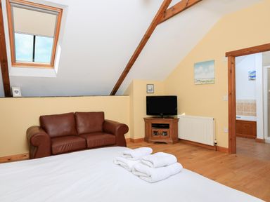 A bedroom with a brown leather sofa a TV on a wooden stand and folded white towels on a bed at Cowslip Cottage in Frogmore