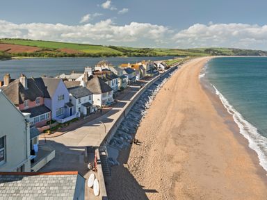 A row of houses along a coastal promenade next to a sandy beach and a body of water with green hills in the background at Cowslip Cottage in Frogmore